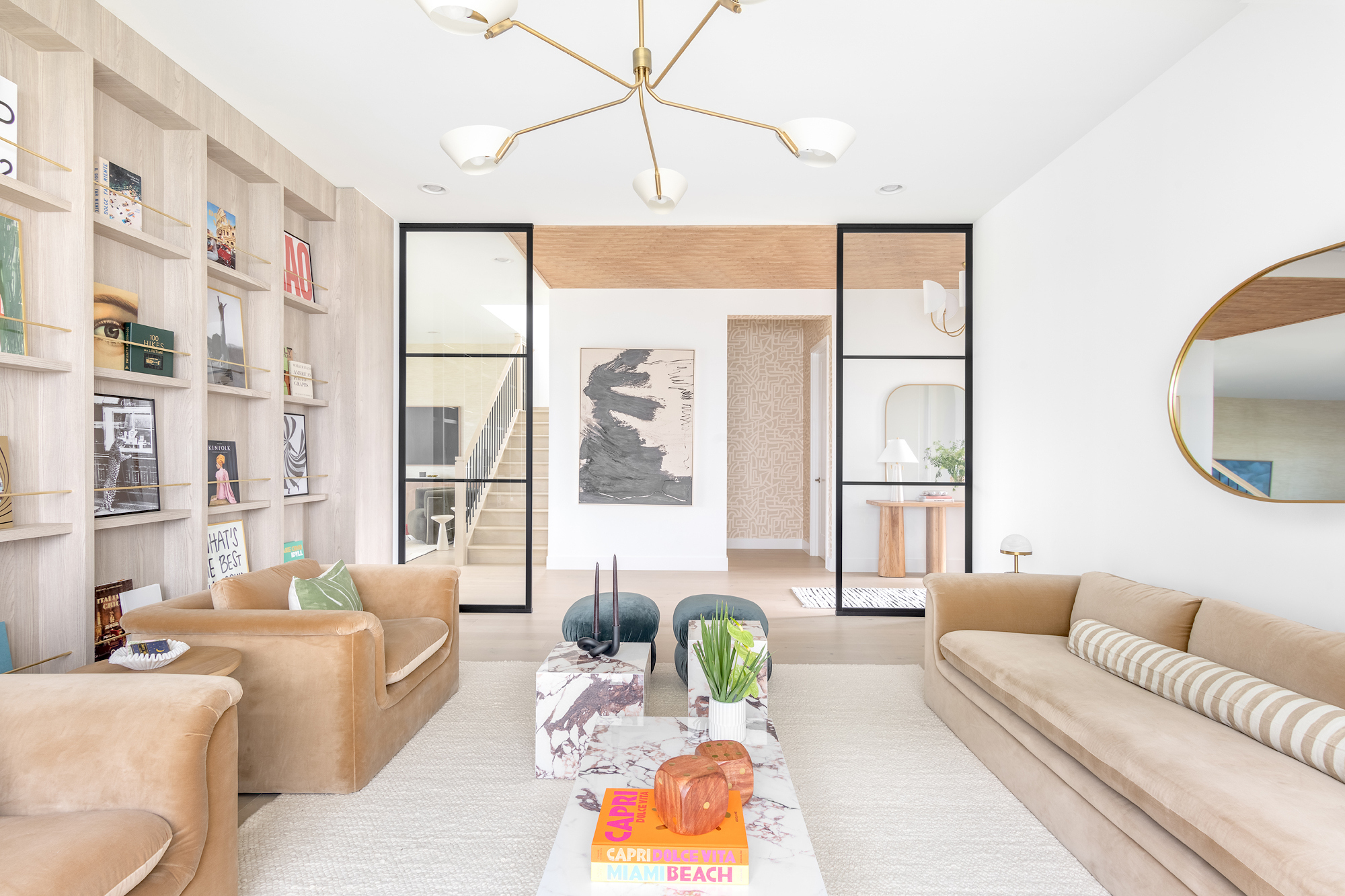 A finished living room with camel velvet sofas, brass details, built-in oak shelving, and Miami coffee-table books