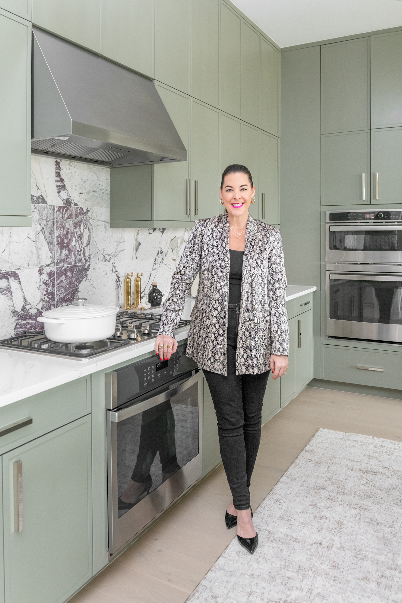 Ann Lopez standing in a renovated kitchen she designed, sage cabinetry and a marble backsplash behind her